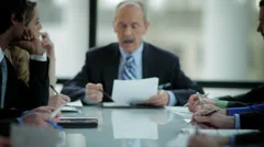 An older businessman leads a meeting at a large table and reads from his paper