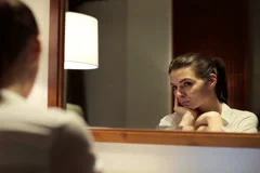 Young pensive woman looking at her reflection in the mirror NTSC