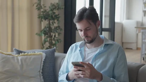 0503 Relaxed Man Browsing Smartphone On Cozy Sofa In Bright Modern Apartment Vidéo 318930475