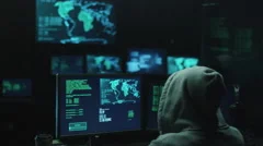 Male hacker in a hood works on a computer with maps and data on display screens