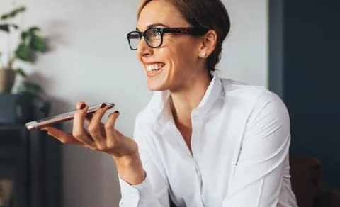 Closeup of smiling business woman talking over mobile phone on loudspeaker. Woman in formal attire holding a mobile phone close to her mouth and looking away. xkwx business, businesswoman, cell phone, close up, collar, entrepreneur, female, formal, g, happy, holding, indoors, lifestyle, looking away, loudspeaker, mobile phone, office, one person, people, phone, sitting, smart, smile, smiling, spectacles, talking, technology, using, white, wireless, worker, working, workplace, young woman