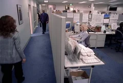 A businessman walks down a long hallway and turns into a large office bustling