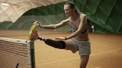 A slender disabled girl stretches her injured leg on the tennis net before the