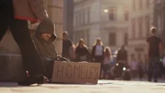 Poor homeless man sitting on street with sign people walk around sunshine