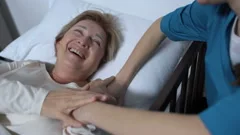 Caring nurse telling jokes to old female patient lying in sickbed rehabilitation