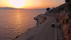 DRONE: Flying behind a big gray car driving along the coastal road at sunrise.