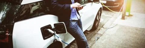 Businessman using mobile phone while charging electric car at charging station xkwx One Person, Mid Adult, Man, Male, Caucasian, Mid Adult Men, Caucasian Ethnicity, Business, Businessman, Executive, Associate, Occupation, Profession, Professional, Skill, Expertise, Entrepreneur, Charging Station, Charging Point, Suit, Smartphone, Mobile Phone, Cellphone, Phone, Wireless Technology, Communication, Connection, Global Communication, Holding, Standing, Charging, Alternative Energy, Renewable Energy, Energy Source, Ecology, Ecological, Efficient, Regenerative, Hybrid, Charger, Car, Electric Car, Charging Port, Electricity, Power Supply, Automobile, Transport, Transportation, Modern, Technology, Millennial, Handsome, Smart, Lifestyle, On The Go, Commuter, Commute, Commuting