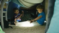 Two boys play with flashlights and walkie talkies inside homemade blanket fort