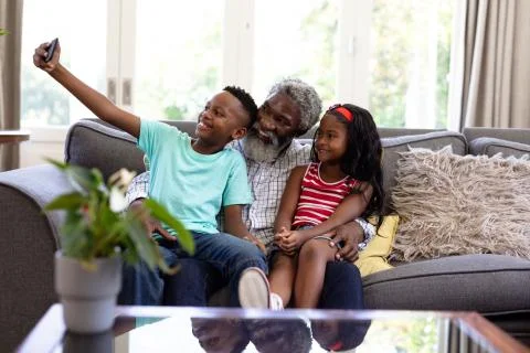 Senior African American man and his grandchildren enjoying their time at home together, sitting on a couch, embracing, smiling, using a smartphone, taking a selfie xkwx Two People, Child, Girl, Female, Mixed-Race, Mid Adult, Man, Male, Black, People, Family, Grandfather, Granddad, Granddaughter, Grandson, Siblings, Brother, Sister, Childhood, Together, Closeness, Interacting, Sitting, Couch, Holding, Using, Phone, Mobile Phone, Smartphone, Connection, Communication, Taking, Photo, Photograph, Selfie, Real People, Home, House, Domestic Life, Lifestyle, Enjoying, Free Time, Leisure, Relaxing, Weekend, Summer, Sunny, Love, Indoors