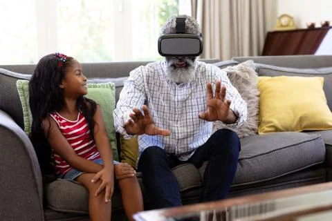 Senior African American man and his granddaughter enjoying their time at home together, sitting on a couch, wearing a VR headset xkwx Two People, Child, Girl, Female, Mixed-Race, Mid Adult, Man, Male, Black, People, Family, Grandfather, Granddad, Granddaughter, Childhood, Together, Closeness, Interacting, Sitting, Couch, Happy, Smiling, Wearing, Using, Vr, Virtual Reality, Headset, Connection, Communication, Real People, Home, House, Domestic Life, Lifestyle, Enjoying, Free Time, Leisure, Relaxing, Weekend, Summer, Sunny, Love, Indoors