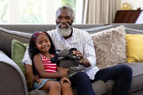 Senior African American man and his granddaughter enjoying their time at home together, sitting on a couch, holding a VR headset, looking at the camera and smiling xkwx Two People, Child, Girl, Female, Mixed-Race, Mid Adult, Man, Male, Black, People, Family, Grandfather, Granddad, Granddaughter, Childhood, Together, Closeness, Interacting, Sitting, Couch, Happy, Smiling, Holding, Vr, Virtual Reality, Headset, Connection, Communication, Real People, Home, House, Domestic Life, Lifestyle, Enjoying, Free Time, Leisure, Relaxing, Weekend, Summer, Sunny, Love, Indoors, Looking At Camera