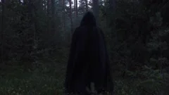 hooded figure in black coat walking away from camera into woods