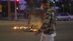 Handheld shot walking past a burning trash container, and people rioting