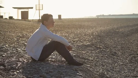 A 16-year- old boy in a white jacket sits on the beach in the wind, watching the Stock Footage 114158892