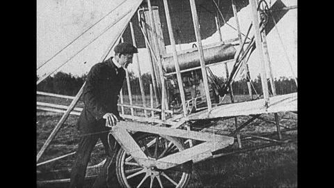 1920s: Man affixes wheel to bottom of wing of Wright brothers' plane. Plane is Stock Footage