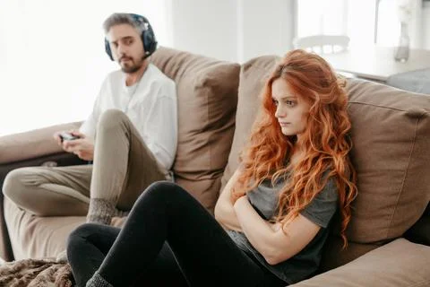 Full body of sad redhead female sitting on comfortable couch near boyfriend playing interesting videogame in light living room at home xkwx couple, videogame, bored, ignore, pastime, leisure, relationship, play, hobby, redhead, sad, together, home, ginger, spend time, upset, boyfriend, girlfriend, long hair, apartment, light, man, woman, flat, red hair, at home, soulmate, entertain, boredom, partner, sofa, living room, couch, headphones, game, weekend, free time, gamepad, chill, problem, gamer, player, frustrate, settee, trouble, disappoint, inside