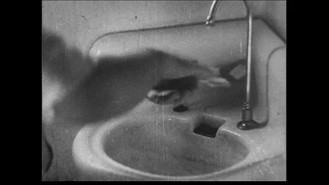1930s: Close up of man drying hands over sink crossfades with medium wide shot Stock Footage