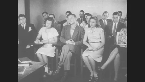 1940s: Boys and girls sit at desks in crowded classroom. Boy in suit folds his Stock Footage
