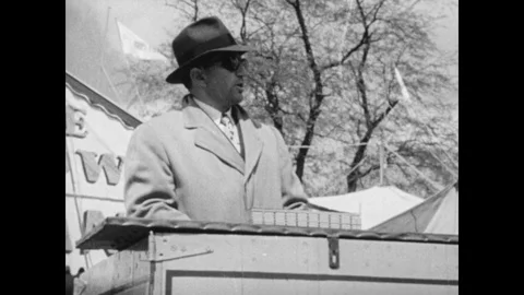 1940s Circus barker speaks at podium near sideshow tent. Crowds of people mill