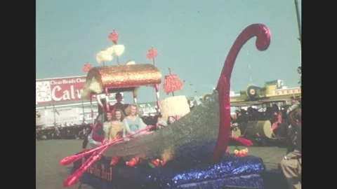 1940s: Women in parade float shaped like boat pass, paddling oars. Woman high on Video stock 77608455