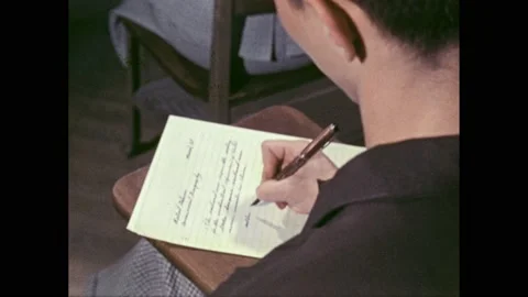 1950s: Boy writes a paragraph on a piece of paper in a classroom. Slate. Boy Vidéo 167182838