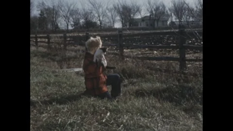 1950s: A child crouches down, exploring the grass near a fence on a clear day, Stock Footage 280856128