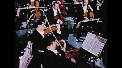 1950s: Conductor leads the orchestra's string section while a majestic horn Stock Footage 98563089