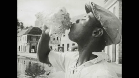 1950s: Man blows into conch shell. Men and women walk through town square. Women Stock Footage 130359120