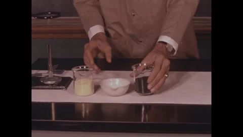 1950s: Man in laboratory sets two glasses of elements on table with burner and Stock Footage 272977117