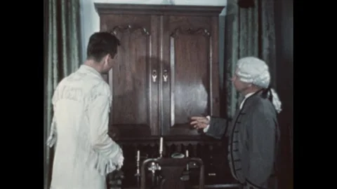 1950s: Man in a powdered wig shows off the woodworking of a cabinet to a man in Stock Footage 291333596