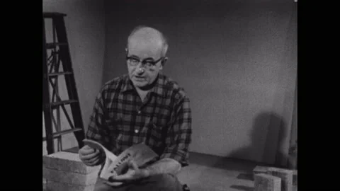 1950s: Man sits on stack of blocks in empty room, talking, holding pamphlet on Stock Footage 151609052