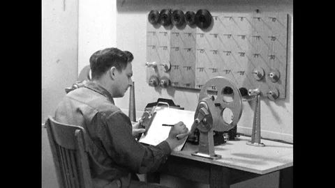 1950s: Man takes notes on clipboard at workbench. Editor places audio tape into Video stock 129569486