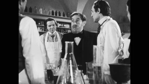 1950s: Men in aprons stand around scientist at laboratory table and speak. Vidéo 143784530