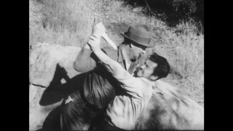 1950s: Scrubby, wild landscape. Two men fight, grapple, man raises knife, other Stock Footage 93325904
