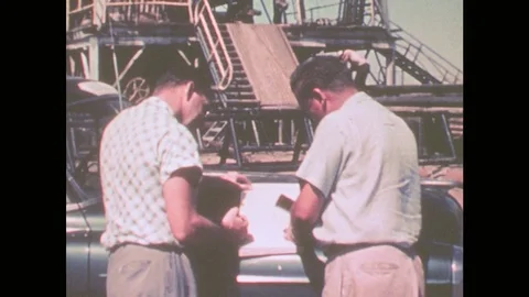 1950s: Two men read a piece of paper together at an oil well site. Two trucks Stock Footage