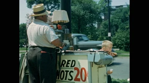 1950s: A young boy buys a hot dog from a vendor with a cart. Boy eats the hot Stock Footage