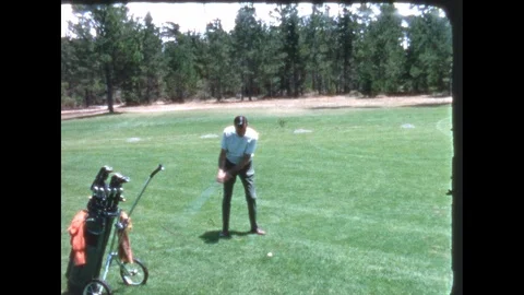 1960s: Golf course, trees, rolling golf bag, man takes practice swing, hits Stock Footage