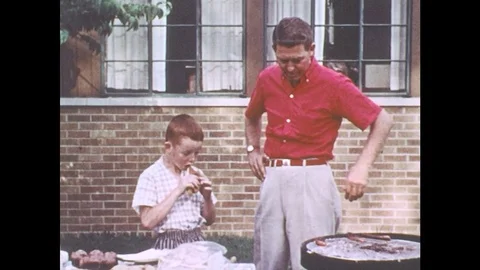 1960s: Man and boy stand next to grill, boy eats hotdog. Field of wheat. Combine Stock Footage 78492792