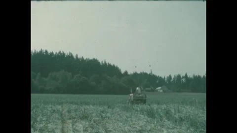 1960s: A man drives a small tractor across a field, pulling a wagon or small Stock Footage 280146196