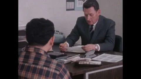 1960s: Man sits at desk with paperwork and interviews another man. Stock Footage 246748897