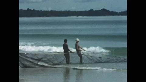 1960s: men pull in fishing nets on beach. Waves on beach. Men by dock. Man on Stock Footage 194187073