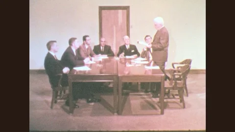 1960s: Men sit at table. Man stands and reads from paper. Men listen and take Stock Footage