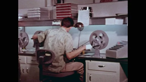 1960s: Office.  People turn knobs to move tape from reel to reel.  Man pushes 스톡 동영상 159489814
