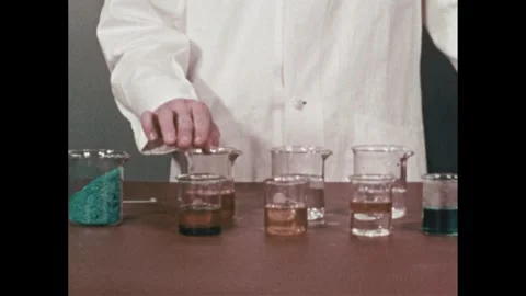 1960s: Table full of various liquids and solutions, scientist's hand points at Video stock 289158624