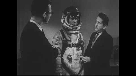 1960s: Two men stand with model in space suit, man describes suit features. Stock Footage