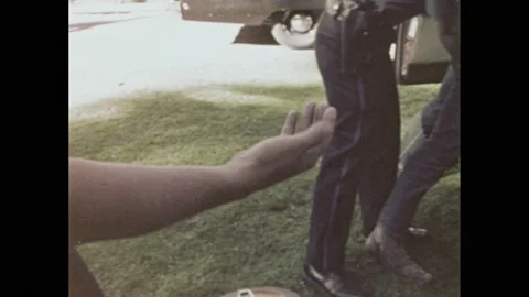 1960s: Two young men and a woman stand with hands on car as a cop frisks the Vídeos de archivo 168386529