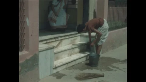 1960s: Young man cleans steps of house.  Woman sits in shade. Stock Footage 248247002