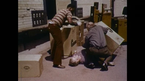 1970s: man collapsed underneath boxes. Men move boxes from casualty. First aider Stock Footage