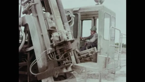 1970s: man in hard hat in cab of drill operates drill and watches as it enters Stock Footage 196949899