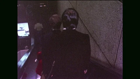 1970s: man rides escalator down at airport. traveller walks to counter for Avis Video stock 90809326